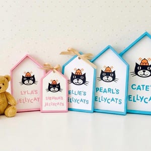 May include: A set of cat-themed name plaques and tags. Each features a black cat illustration wearing a hat, with names such as "Lyla's Jellycat" and "Cate's Ellycats". The plaques have pink and blue frames. A teddy bear is also present.