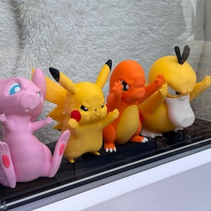 May include: A set of four small plastic character figurines arranged in a row. The figures include a pink Mew, a yellow Pikachu, an orange Charmander, and a yellow Psyduck. Each character is depicted with a focused, determined facial expression.