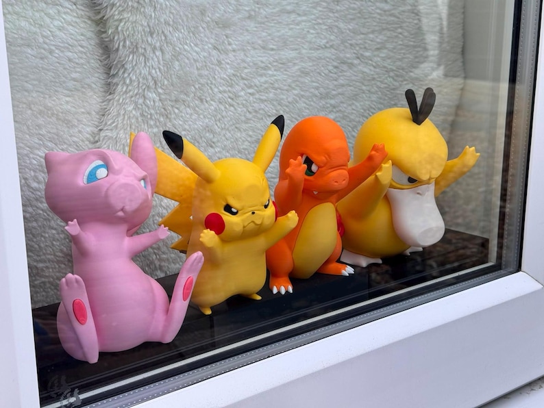 May include: A set of four small plastic character figurines arranged in a row. The figures include a pink Mew, a yellow Pikachu, an orange Charmander, and a yellow Psyduck. Each character is depicted with a focused, determined facial expression.