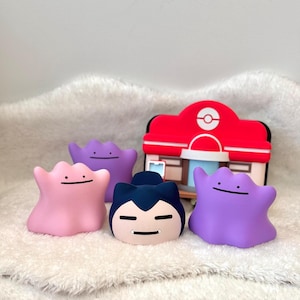 May include: A collection of four small character figurines including three amorphous blob-like figures in pink and purple shades, one dark blue and cream-colored figure with ears, and a red and white building structure with a circular logo.