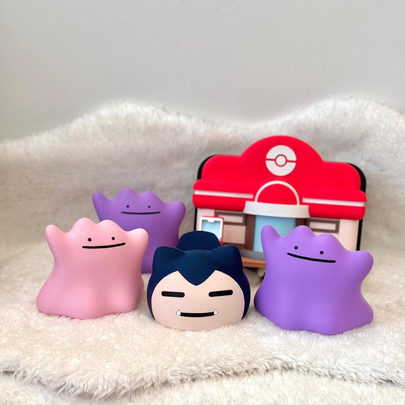 May include: A collection of four small character figurines including three amorphous blob-like figures in pink and purple shades, one dark blue and cream-colored figure with ears, and a red and white building structure with a circular logo.