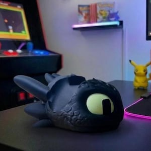 May include: A black and grey dragon-shaped desk tidy with a large yellow eye. The organiser is on a black desk, with an arcade machine and a Pikachu figure in the background. The dragon has a textured, scaly appearance.