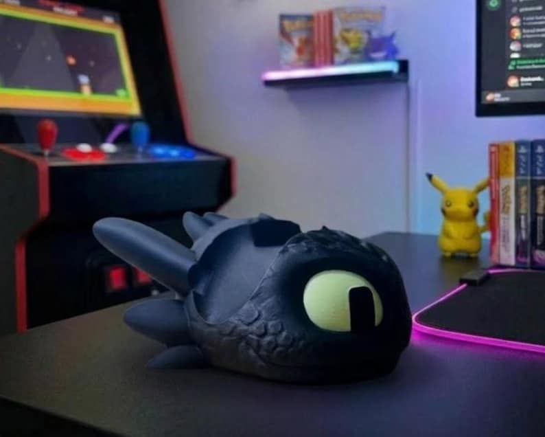 May include: A black and grey dragon-shaped desk tidy with a large yellow eye. The organiser is on a black desk, with an arcade machine and a Pikachu figure in the background. The dragon has a textured, scaly appearance.
