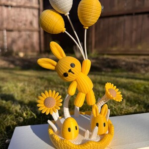 May include: A yellow 3D-printed Easter bunny figurine holding three balloons, with a basket of flowers and an egg. The bunny is in mid-air, with a happy expression. The figurine is on a white surface.