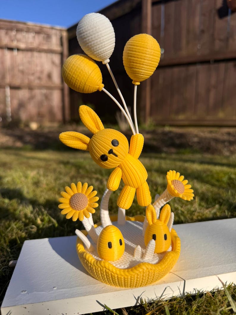May include: A yellow 3D-printed Easter bunny figurine holding three balloons, with a basket of flowers and an egg. The bunny is in mid-air, with a happy expression. The figurine is on a white surface.