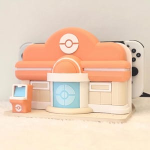 May include: A peach and cream colored gaming console dock shaped like a building with a central blue door, a circular emblem on the roof, and a small matching kiosk on the left side. A white gaming console is inserted into the back of the structure.