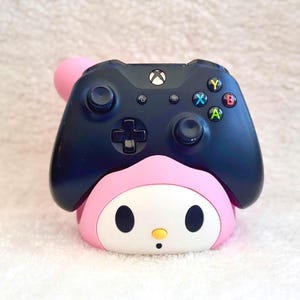 Peut inclure: Une manette de jeu vidéo sans fil noire reposant sur un support à l'effigie d'un personnage rose et blanc. La manette comprend un bouton central avec le logo Xbox, deux joysticks, une croix directionnelle et quatre boutons d'action colorés.