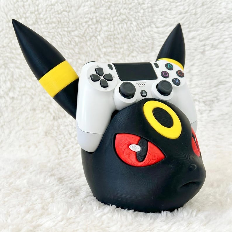 May include: A black and yellow controller stand shaped like a creature head with pointed ears, featuring a white game controller resting on top. The stand has bright red eyes and a yellow circular forehead marking, designed to hold gaming hardware.