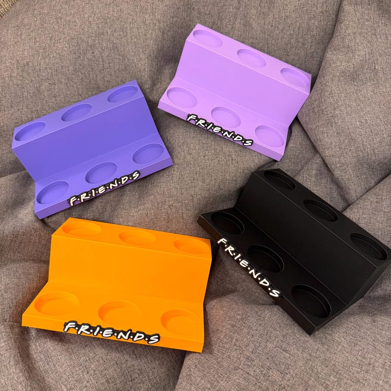 May include: Four colorful, angled display stands in purple, orange, black, and lavender. Each stand has six circular cutouts and the word "FRIENDS" in white, stylized text. The stands are made of a solid material and are arranged on a gray surface.
