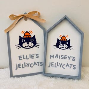 Puede incluir: Dos letreros en forma de casa con marcos grises y centros blancos. Cada uno presenta una ilustraci&oacute;n de un gato negro con un sombrero naranja y l&iacute;neas de movimiento naranjas. El letrero izquierdo tiene una cinta beige y dice "ELLIE'S JELLYCATS", mientras que el derecho dice "MAISEY'S JELLYCATS".