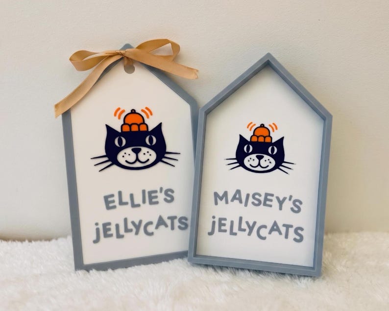 Puede incluir: Dos letreros en forma de casa con marcos grises y centros blancos. Cada uno presenta una ilustraci&oacute;n de un gato negro con un sombrero naranja y l&iacute;neas de movimiento naranjas. El letrero izquierdo tiene una cinta beige y dice "ELLIE'S JELLYCATS", mientras que el derecho dice "MAISEY'S JELLYCATS".