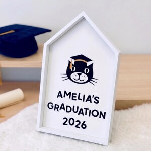 May include: A white, house-shaped sign with the text "AMELIA'S GRADUATION 2026" and a cat wearing a graduation cap. A blue graduation cap with a gold tassel is visible in the background.