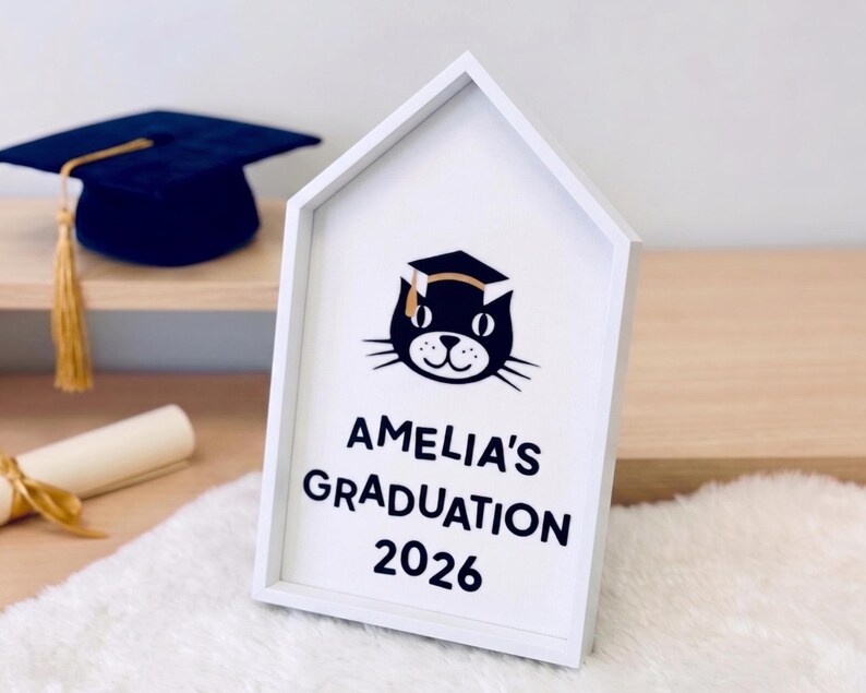 May include: A white, house-shaped sign with the text "AMELIA'S GRADUATION 2026" and a cat wearing a graduation cap. A blue graduation cap with a gold tassel is visible in the background.