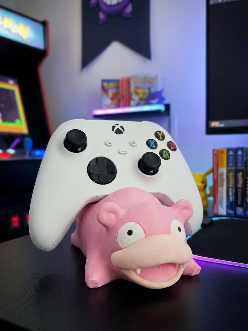 Pode incluir: Um controle Xbox branco repousa sobre um suporte rosa em forma de Slowpoke. O suporte apresenta detalhes, com olhos, boca e orelhas vis&iacute;veis. O fundo inclui uma configura&ccedil;&atilde;o de jogos.