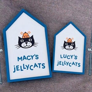 May include: Two house-shaped signs with a blue border and white interior. Each sign features a black cat illustration wearing a hat, with the text "MACY'S JELLYCATS" and "LUCY'S JELLYCATS". The larger sign measures 26.2 cm in height.