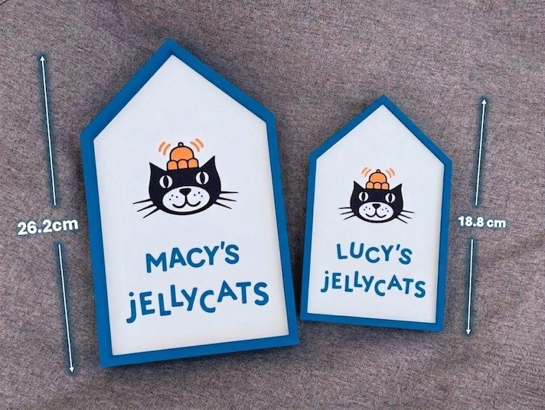 May include: Two house-shaped signs with a blue border and white interior. Each sign features a black cat illustration wearing a hat, with the text "MACY'S JELLYCATS" and "LUCY'S JELLYCATS". The larger sign measures 26.2 cm in height.