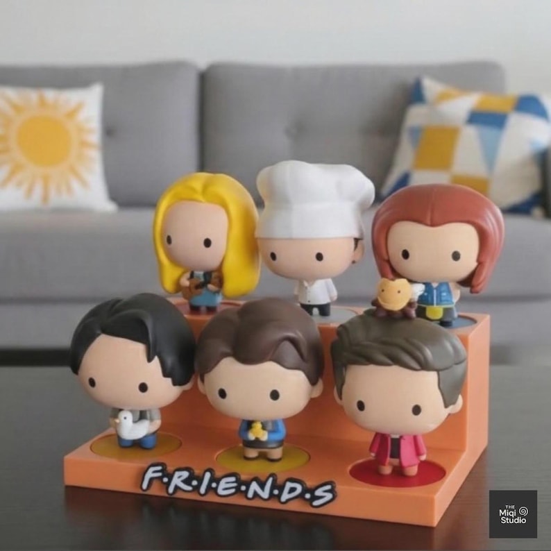 May include: A display of seven cartoon figurines of the Friends television show characters. The figurines are on an orange display stand with the show's logo. The characters have large heads and are wearing outfits that represent their roles in the show.