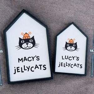 May include: Two house-shaped signs with black frames. The larger sign is 26.2 cm tall and the smaller is 18.8 cm tall. Each sign features a black cat illustration wearing a hat and the text "MACY'S JELLYCATS" and "LUCY'S JELLYCATS".