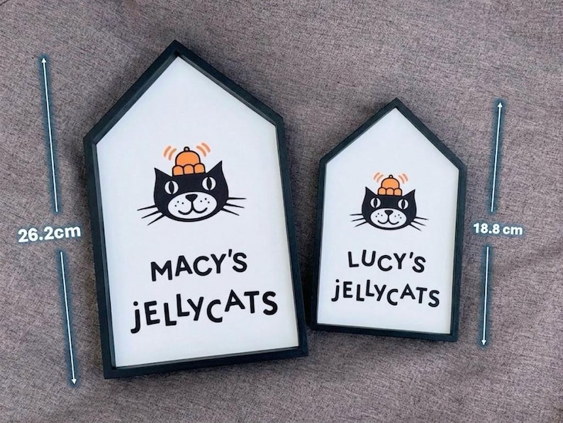 May include: Two house-shaped signs with black frames. The larger sign is 26.2 cm tall and the smaller is 18.8 cm tall. Each sign features a black cat illustration wearing a hat and the text "MACY'S JELLYCATS" and "LUCY'S JELLYCATS".