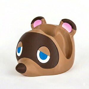 May include: A brown and tan desk organizer shaped like a cartoon character's head, with large eyes and ears. The eyes are white with blue accents. The ears have pink inner details. The organizer is designed to hold small items.
