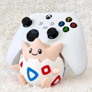 May include: A white wireless video game controller positioned behind a small, round, light-coloured creature figure. The figure has a smooth, pale peach-coloured head with a simple face and a white egg-shaped body featuring red and blue geometric shapes.