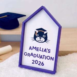 May include: A purple framed sign with the text "AMELIA'S GRADUATION 2026" and a cat wearing a graduation cap. A navy blue graduation cap with a gold tassel is in the background.