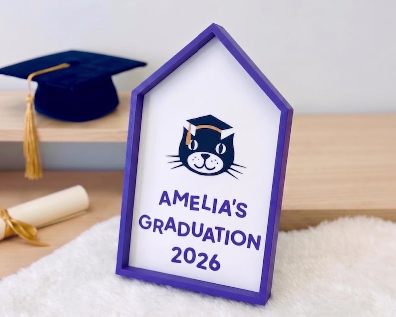 May include: A purple framed sign with the text "AMELIA'S GRADUATION 2026" and a cat wearing a graduation cap. A navy blue graduation cap with a gold tassel is in the background.