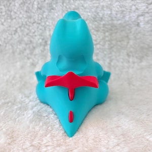 May include: A turquoise figurine with a red accent on its back and tail. The figurine has a smooth, rounded shape and is set against a white, textured background. The figurine is a stylized representation of a creature.