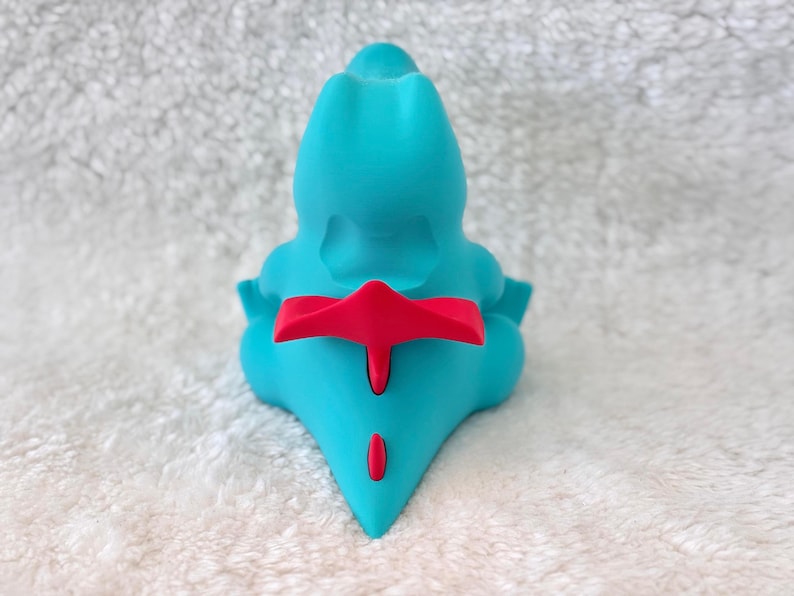 May include: A turquoise figurine with a red accent on its back and tail. The figurine has a smooth, rounded shape and is set against a white, textured background. The figurine is a stylized representation of a creature.