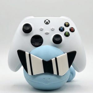 Puede incluir: Un mando blanco de Xbox descansa sobre una figura azul claro que lleva gafas de sol a rayas blancas y negras. El mando tiene joysticks negros y botones de colores. La figura tiene una forma suave y redondeada.