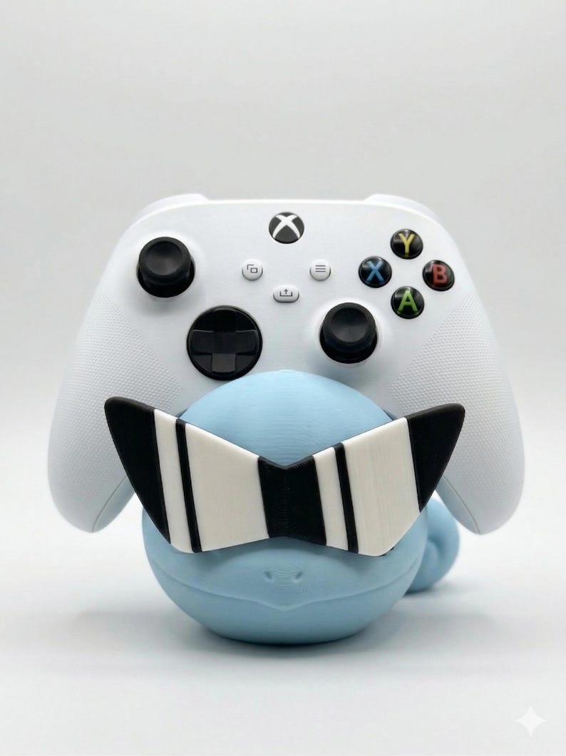 Puede incluir: Un mando blanco de Xbox descansa sobre una figura azul claro que lleva gafas de sol a rayas blancas y negras. El mando tiene joysticks negros y botones de colores. La figura tiene una forma suave y redondeada.