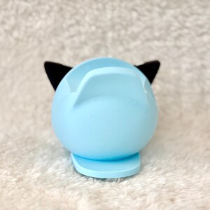 Puede incluir: Un soporte para tel&eacute;fono con forma de gato azul claro con orejas negras. El soporte tiene una base redondeada y una parte superior curvada para sostener un tel&eacute;fono. El dise&ntilde;o es simple y moderno, adecuado para un escritorio o mesita de noche.