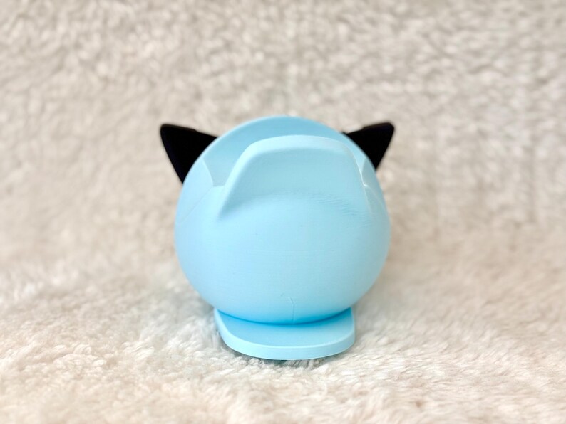Puede incluir: Un soporte para tel&eacute;fono con forma de gato azul claro con orejas negras. El soporte tiene una base redondeada y una parte superior curvada para sostener un tel&eacute;fono. El dise&ntilde;o es simple y moderno, adecuado para un escritorio o mesita de noche.
