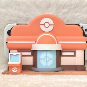 May include: A 3D-printed model of a building with an orange roof and a white and blue circular design. The model has a brown base and a small orange kiosk. A white and black game console is visible behind the model.
