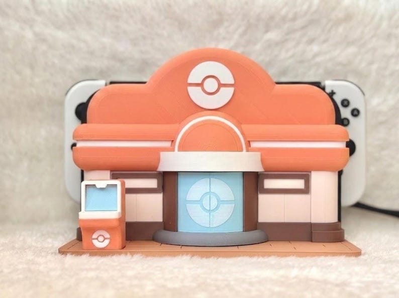 May include: A 3D-printed model of a building with an orange roof and a white and blue circular design. The model has a brown base and a small orange kiosk. A white and black game console is visible behind the model.