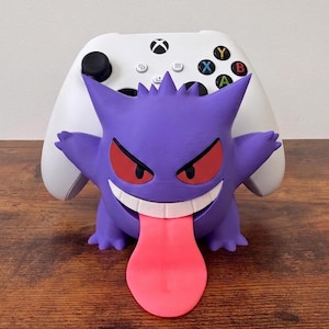May include: A purple Gengar character controller stand featuring red eyes, a wide white grin, and a long pink tongue, holding a white Xbox wireless controller with black thumbsticks and multicolored A, B, X, and Y action buttons on the front.