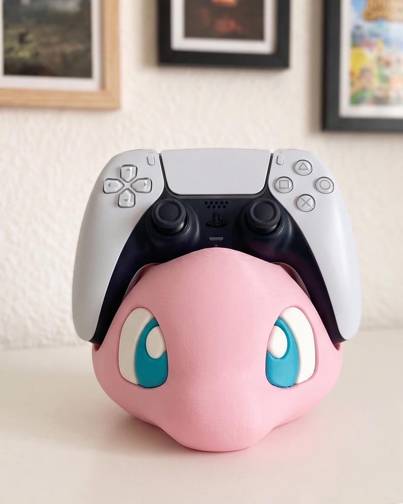 May include: A pink, 3D-printed character head with large blue eyes, holding a white and black PlayStation 5 controller. The controller is resting on top of the character's head. The character has a smooth, matte finish.