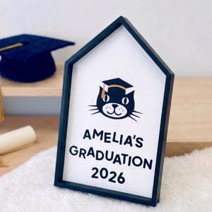 May include: A black framed sign with the text "AMELIA'S GRADUATION 2026" and a cat wearing a graduation cap. A navy blue graduation cap with a gold tassel and a rolled diploma with a gold ribbon are also visible.