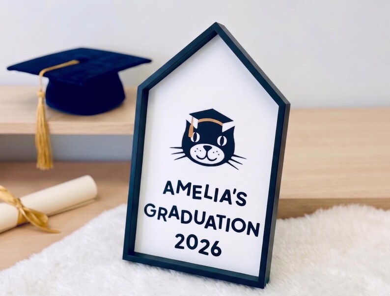 May include: A black framed sign with the text "AMELIA'S GRADUATION 2026" and a cat wearing a graduation cap. A navy blue graduation cap with a gold tassel and a rolled diploma with a gold ribbon are also visible.