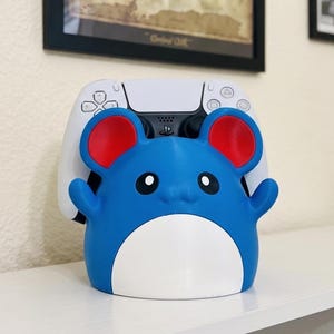 May include: A blue and white controller holder shaped like a cartoon mouse with red ears. The controller holder is designed to hold a white PlayStation 5 controller. The holder is on a white shelf.