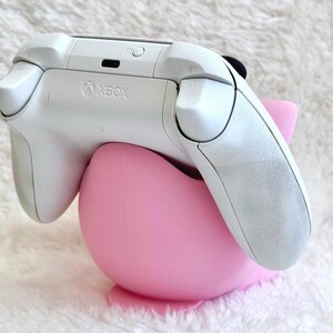 Peut inclure: Une manette sans fil Xbox blanche pos&eacute;e sur un support en plastique rose clair, lisse et arrondi. La manette pr&eacute;sente le logo Xbox au centre sup&eacute;rieur, avec les boutons de tranche et les g&acirc;chettes visibles. Le support est incurv&eacute;.