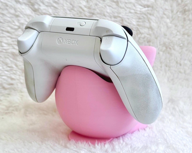 Peut inclure: Une manette sans fil Xbox blanche pos&eacute;e sur un support en plastique rose clair, lisse et arrondi. La manette pr&eacute;sente le logo Xbox au centre sup&eacute;rieur, avec les boutons de tranche et les g&acirc;chettes visibles. Le support est incurv&eacute;.