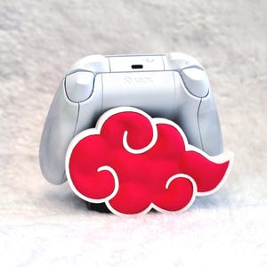 May include: A white Xbox game controller stand featuring a prominent red cloud design with a white outline. The controller holder is shaped like a stylized cloud, providing a secure base for the white gaming controller resting behind it.