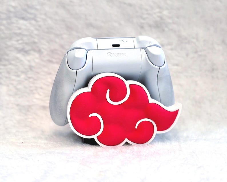 May include: A white Xbox game controller stand featuring a prominent red cloud design with a white outline. The controller holder is shaped like a stylized cloud, providing a secure base for the white gaming controller resting behind it.