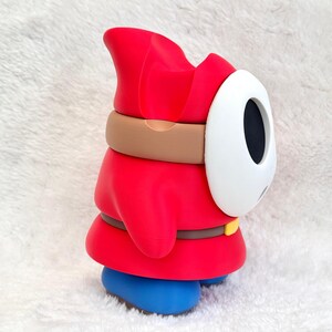 May include: A red figurine featuring a white oval mask with black eye and mouth openings. The character wears a red hooded robe, a brown belt with a yellow buckle, and blue shoes. The design is smooth and matte, capturing the side profile of the character.