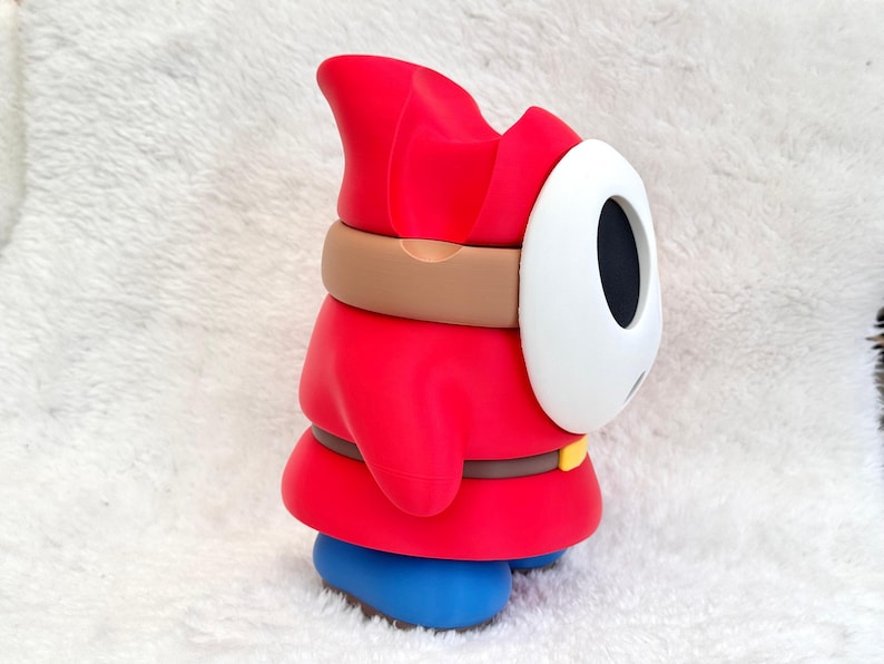 May include: A red figurine featuring a white oval mask with black eye and mouth openings. The character wears a red hooded robe, a brown belt with a yellow buckle, and blue shoes. The design is smooth and matte, capturing the side profile of the character.