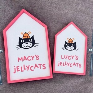 May include: Two house-shaped signs with pink frames. The larger sign is 26.2 cm tall, and the smaller is 18.8 cm. Each sign features a black cat illustration with a pumpkin on its head and the text "MACY'S JELLYCATS" and "LUCY'S JELLYCATS".