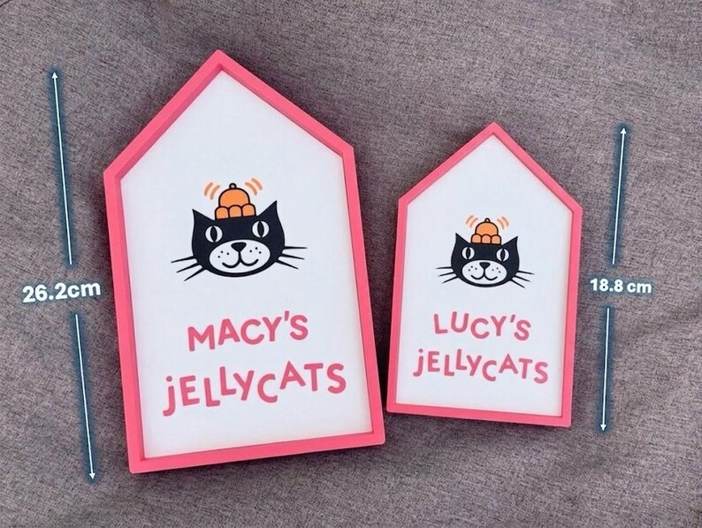 May include: Two house-shaped signs with pink frames. The larger sign is 26.2 cm tall, and the smaller is 18.8 cm. Each sign features a black cat illustration with a pumpkin on its head and the text "MACY'S JELLYCATS" and "LUCY'S JELLYCATS".