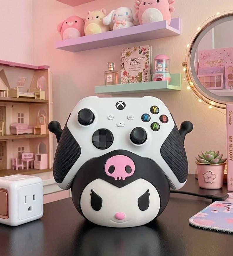 May include: A white Xbox controller sits atop a black and white Kuromi-themed base. The controller features black thumbsticks and coloured buttons. A pink skull is centered on the base. Shelves with plush toys and a dollhouse are in the background.