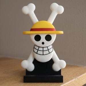 May include: A white and black figurine of a cartoon skull with a yellow hat and red band, and two white crossbones. The figurine is on a black base and sits on a wooden surface.